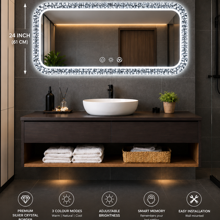 A rectangular LED-backlit bathroom mirror with a sparkling silver crystal frame, mounted on a dark gray tiled wall above a modern dark wood vanity with a white vessel sink. The image includes dimension lines indicating the mirror is 48 inches by 24 inches, and feature icons at the bottom describing its properties.