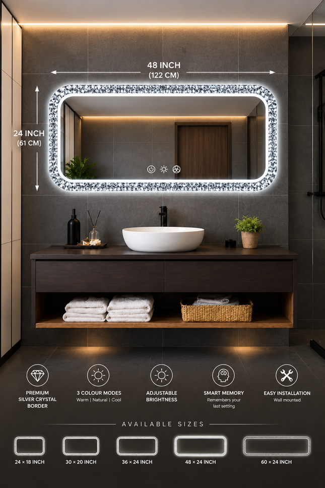 A rectangular LED-backlit bathroom mirror with a sparkling silver crystal frame, mounted on a dark gray tiled wall above a modern dark wood vanity with a white vessel sink. The image includes dimension lines indicating the mirror is 48 inches by 24 inches, and feature icons at the bottom describing its properties.